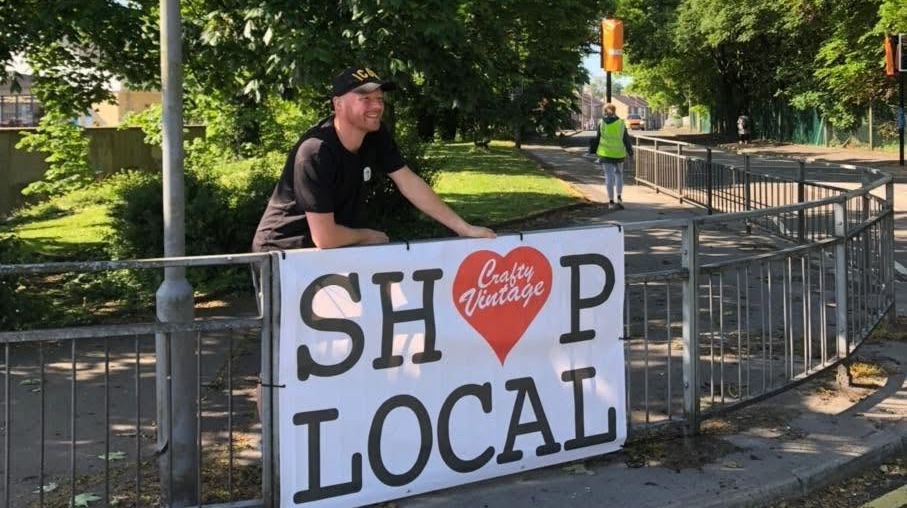 Dan Johnson leaning on a railing behind a large Shop Local banner on a tree-lined street